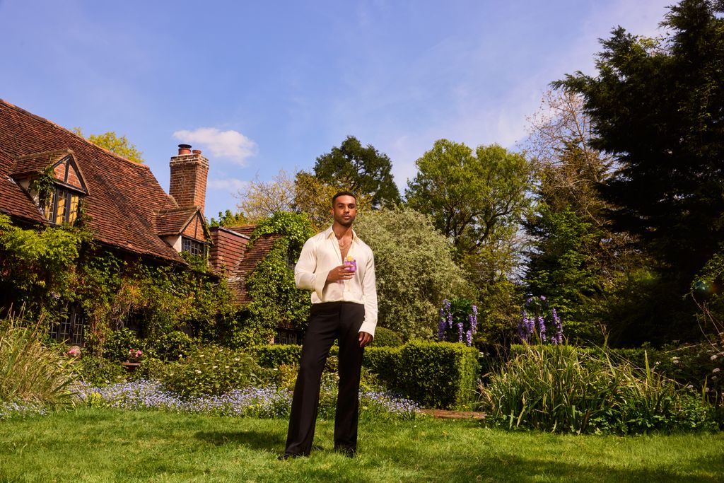 Lucien Laviscount holding a drink in front of a cottage