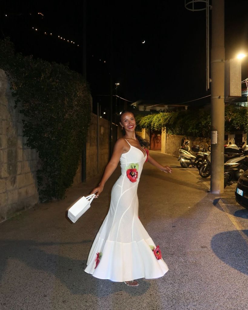 Maya Jama poses under a lush canopy of lemon trees, likely in an al fresco dining setting, possibly in Italy or the South of France. She wears a sheer, white crochet maxi dress that hugs her curves elegantly, adorned with large, vibrant red poppy appliqués. The dress features thin spaghetti straps and a plunging neckline, striking a perfect balance between sultry and sophisticated. She accessorizes with a structured white mini handbag and white sandals, her deep red lipstick echoing the floral embellishments on her dress. Her curly hair is styled to one side, revealing her glowing complexion and gold hoop earrings. She stands on a beautifully tiled floor, with warm ambient lighting adding to the romantic, Mediterranean vibe.